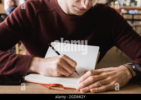 A young man sits in cafe, drinks coffee and makes notes and sketches in notebook Stock Photo