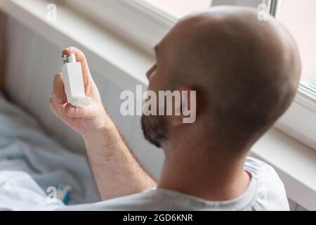 A bald, bearded man holds an inhaler in his hand and lying in bed. Top view, from the back. The concept of World Asthma Day. Stock Photo
