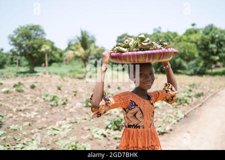 Smart african girl smiling into the camera, carrying a plate with dried leaves on her head, on her way to the traditional medicine market Stock Photo