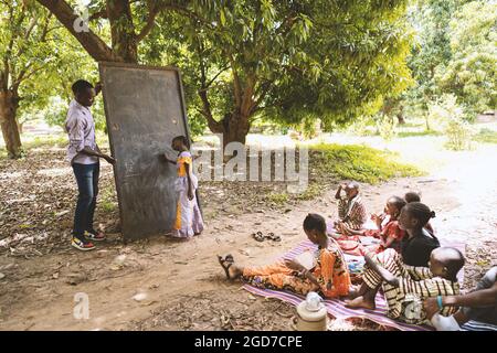 Little African girl at the blackboard mentally calculating a sum with her head tilted, under the attentive gaze of the teacher, with group of children Stock Photo