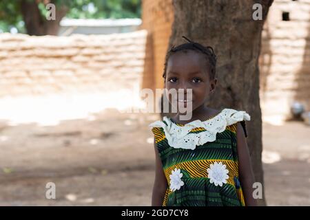 Adorable little black African girl standing before a tree in the courtyard of her home in a remote African village; poverty concept Stock Photo