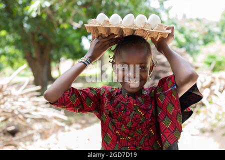 In this image, a pretty little African girl with earrings and bangles in a sophisticatedly cut dress is posing with a carton full of eggs on her head, Stock Photo