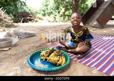 Gentle little black African girl sitting on a coloured mat in the middle of a rural courtyard, offering a banana from a big plate full of fruits Stock Photo