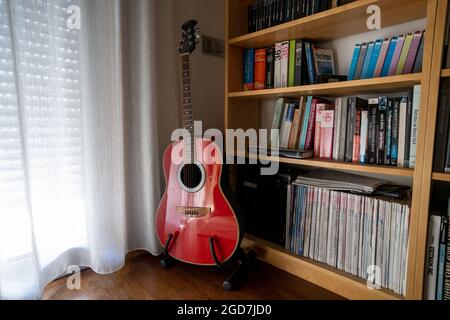 Red acoustic guitar on a stand at home, next to a bookcase full of books and magazines Stock Photo