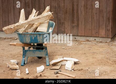 Wheelbarrow full of old building materials before it is wheeled away in preparation for a new concrete patio next to a building structure. Stock Photo