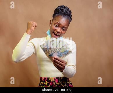 Happy cute African lady with nose mask, holding multiple Naira notes, cash, Nigeria currency or money used for transactions in her hands Stock Photo