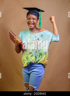 An African Nigerian lady or graduand, wearing a graduation cap and glasses, carrying books as she expresses joy due to success in her school, educatio Stock Photo