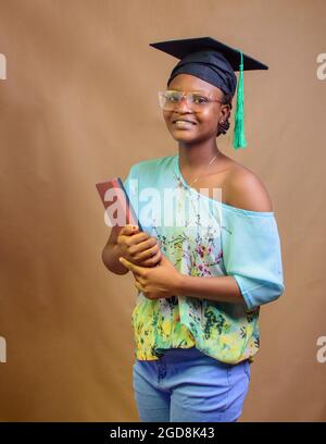 An African Nigerian lady or graduand, wearing a graduation cap and glasses, carrying books as she expresses joy due to success in her school, educatio Stock Photo