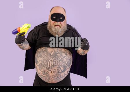 Funny evil obese man in carnival costume holds toy blaster posing on purple background Stock Photo