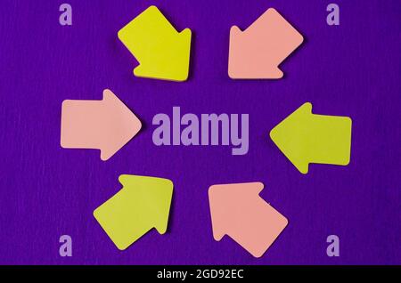 Paper sticky notes in shape of arrow on lilac background. Pink and yellow arrows around the empty space in the center. Top view Stock Photo