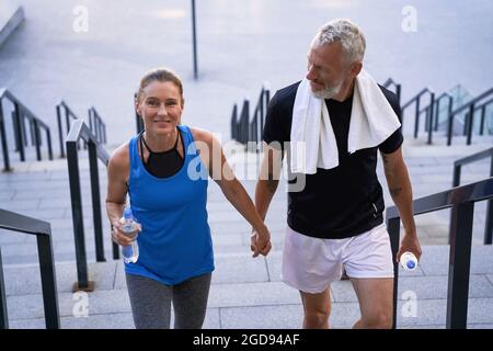 Attractive middle aged couple of sportive man and woman looking happy, holding hands while walking up the stairs after training together in the city Stock Photo