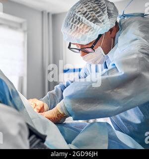 Male plastic surgeon in glasses performing abdominoplasty or tummy tuck surgery in operating room. Man doctor wearing surgical uniform and medical face mask while doing abdominal plastic surgery. Stock Photo