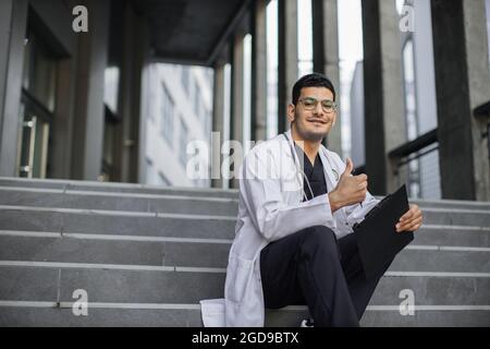 Close up horizontal shot of smiling Indian man physician, wearing scrubs and medical coat, sitting with clipboard on modern stairs outdoors and showing like to camera. Copy space Stock Photo