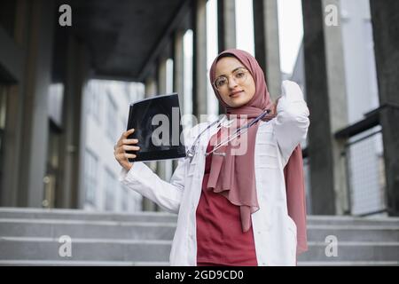 Outdoor portrait of muslim arab female medical doctor holding xray scan for checking disease progress, copy space. Young woman physician in headscarf holding x-ray, standing outside modern clinic Stock Photo