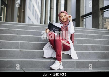 Pretty muslim asian female medical doctor, wearing hijab, red scrubs and white coat, sitting on modern gray stairs outside, holding xray tomography scan and adjusting eyeglasses Stock Photo
