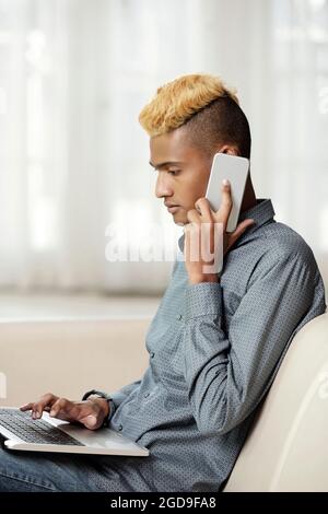 Serious young software developer working on laptop at home and talking on phone with client and discussing changes he needs to do Stock Photo