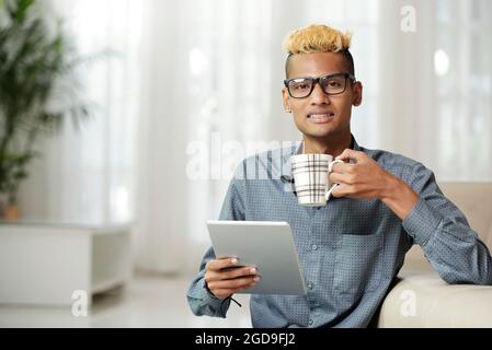 Portrait of intelligent young man reading e-book on programming and software development and drinking hot coffee Stock Photo