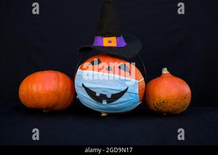 Halloween decorations in the new reality of the COVID-19 pandemic Three pumpkins, one of them wearing a witch hat and a medical mask with a scary smil Stock Photo