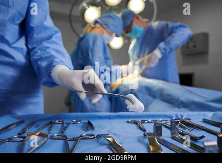Close up of doctor hand holding forceps with tampon while surgeon and assistant performing plastic surgery on blurred background. Medical team using v Stock Photo