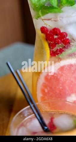 Grapefruit lemonade with mint, lemon, currants and ice in a decanter and a glass with black plastic tubes on a background of fruit. Cooling, refreshing, fruit, summer drink. Vertical close up Stock Photo