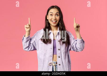 Enthusiastic young pretty girl looking and pointing fingers up with happy smile, see promo, interested in taking part event, party, found great online Stock Photo