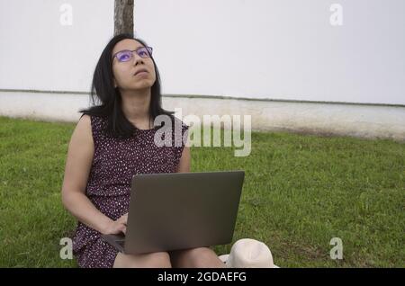 Thoughtful young Hispanic female with glasses sitting on the grass working with the laptop Stock Photo