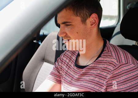 Happy smiling caucasian young man sitting in car. 20s guy looking from open window. Silver grey gray car. Attractive brunette men in vehicle. Buyer or Stock Photo