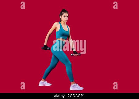 Young slim smiling woman in sports clothes taking a step forward with dumbbells holding un her hands doing fitness. Stock Photo