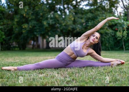 Young woman in sportswear stretching in split pose at garden on grass, outdoors Stock Photo