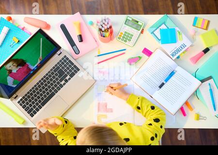 Back to school. Upper view of modern girl with workbooks, stationary, laptop and textbook homeschooling and video chatting at white table at home. Stock Photo