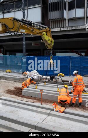 Team of workmen wearing orange high-viz overalls on a construction site work at laying the new track foundations for the update to the Midland Metro tram public transport system in the city centre along Corporation Street on 3rd August 2021 in Birmingham, United Kingdom. The original tracks are being pulled up and relaid, while a new line is also under construction and due to open later in the year. The Midland Metro is a light-rail tram line in the county of West Midlands, England, operating between the cities of Birmingham and Wolverhampton via the towns of West Bromwich and Wednesbury. The Stock Photo