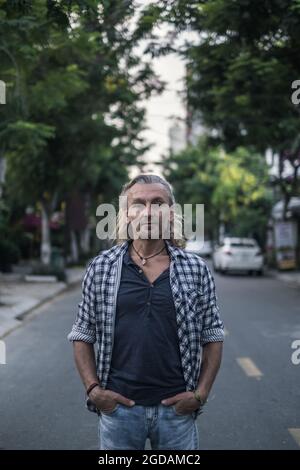 Portrait of elderly handsome man in shirt and jeans standing on the street in town. hands ib his pockets. Long grey hair. Portrait. High quality photo Stock Photo
