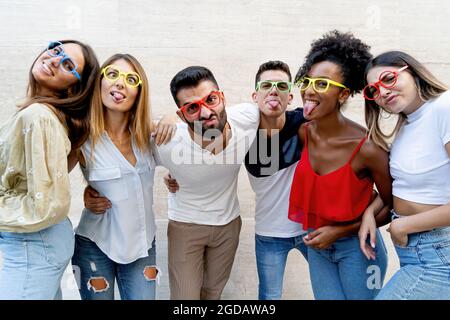 Interracial gen z young people having fun making weird faces and wearing colored glasses. Stock Photo