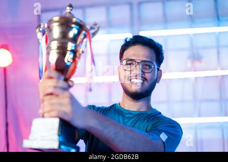 Cheerful happy smiling gamer with headphones celebrating byholding winning trophy at esports gaming tournament stage Stock Photo