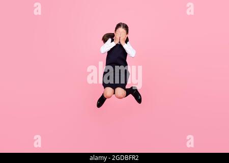 Full length body size photo schoolgirl jumping up covering face with hands isolated pastel pink color background