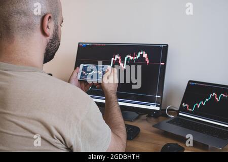 The young man is studying the market charts on both the phone and the computer screen. Stock Photo