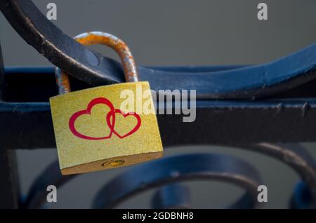 Golden padlock with two red hearts and rusty bracket hangs on black rounded swirls of bridge fence across river on a blurred background of water. Conc Stock Photo