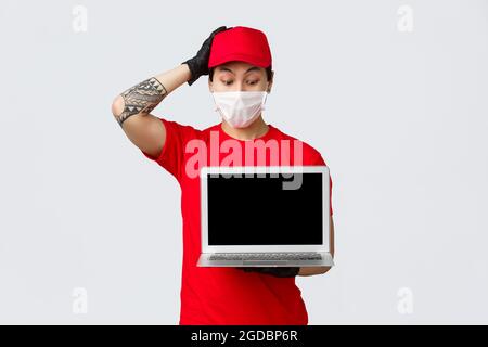 Confused and indecisive asian delivery guy holding laptop, looking puzzled at computer screen, scratch head clueless, wearing courier service uniform Stock Photo