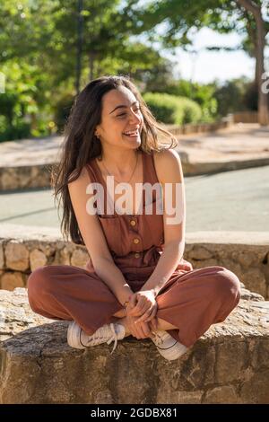 Happy smiling young woman sitting cross-legged. She is sitting on a stone bench in a park. Caucasian with dark hair. She is wearing a red dress. Javea Stock Photo