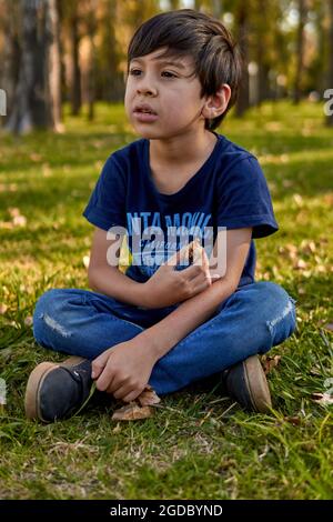 thoughtful brown boy eating while sitting in the middle of the forest at autumn. blurred background. vertical Stock Photo