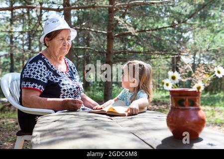 Granddaughter smell flowers with her senior granny, women sitting in forest and talking Stock Photo