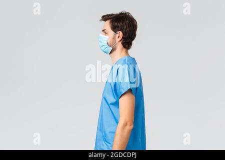 Covid-19, quarantine, hospitals and healthcare workers concept. Profile portrait of young doctor fighting coronavirus, standing grey background in Stock Photo