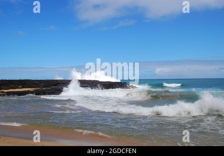 Beautiful blue sky and turquoise water surround this outcropping of black lava rock on the island of Kauai, Hawaii.  Waves crash against rocks. Stock Photo