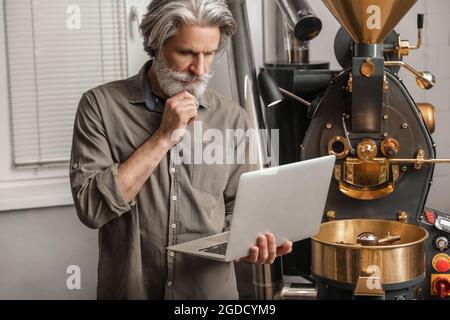 Senior man with laptop using modern coffee roaster Stock Photo