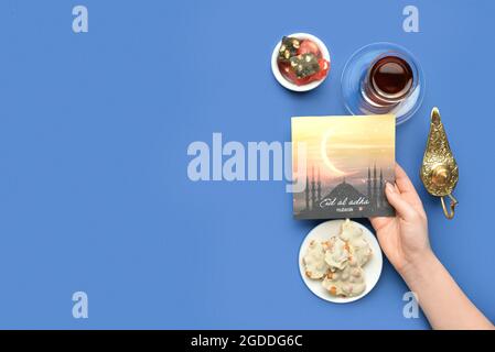 Female hand with greeting card for Eid al-Adha (Feast of the Sacrifice), Turkish sweets and tea on color background Stock Photo