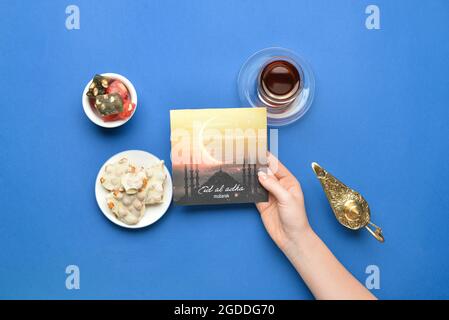 Female hand with greeting card for Eid al-Adha (Feast of the Sacrifice), Turkish sweets and tea on color background Stock Photo