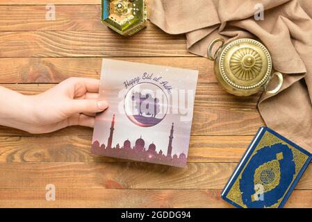 Female hand with greeting card for Eid al-Adha (Feast of the Sacrifice), Quran and lantern on wooden background Stock Photo