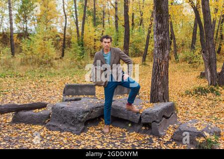 Tall handsome man dressed in a brown jacket posing with a bag near the rocks in autumn forest Stock Photo