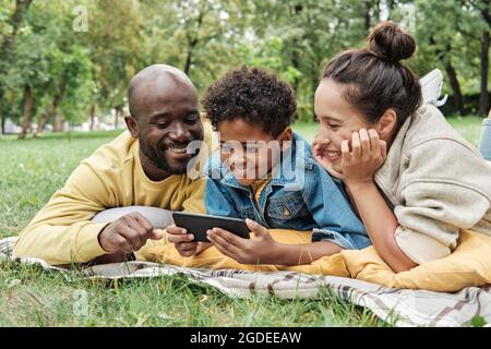 African happy family lying on the grass in the park watching photos on mobile phone and laughing Stock Photo