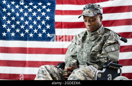 Patriotic Soldier Sitting In Wheel Chair Against American Flag Stock Photo